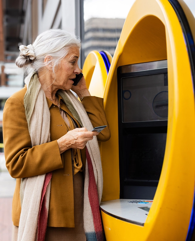 Mujer adulta usando un cajero automático mientras sostiene su tarjeta y habla por teléfono, ejemplo de autonomía digital en la silver economy.