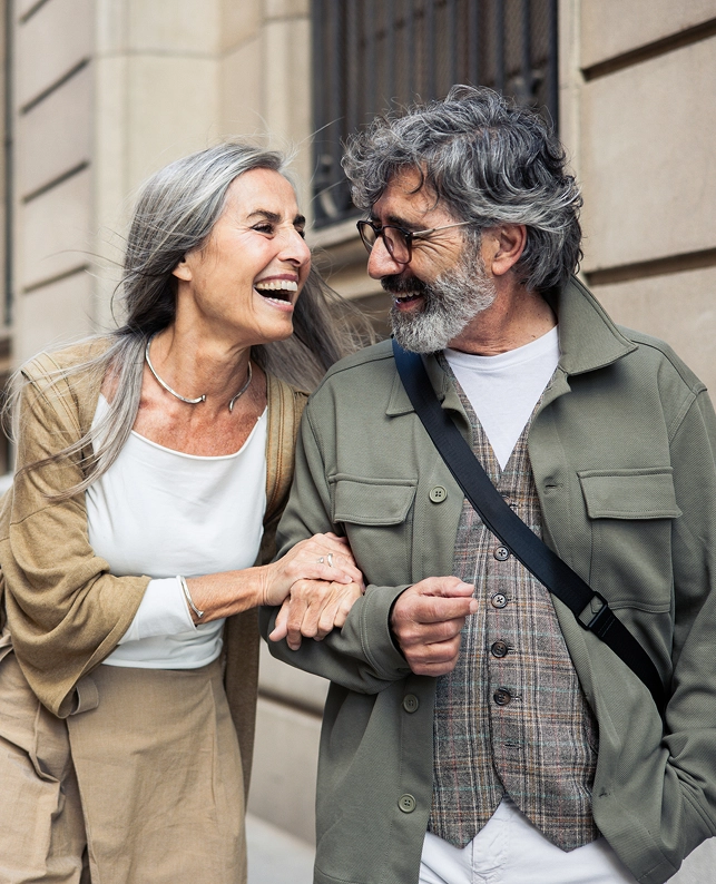 Pareja mayor caminando por la calle mientras se miran y sonríen, representación de conexión afectiva y envejecimiento activo.